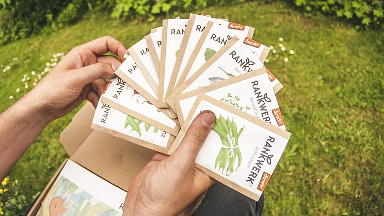 Person hält mehrere Rankwerk-Saatguttüten in der Hand, umgeben von grüner Wiese im Hintergrund.
