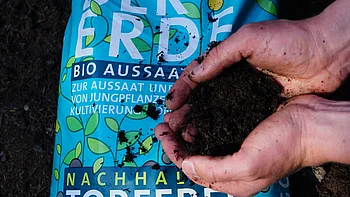 Nahaufnahme eines Paares von Händen, die Erde halten, im Hintergrund ist ein Beutel mit Dünger zu sehen