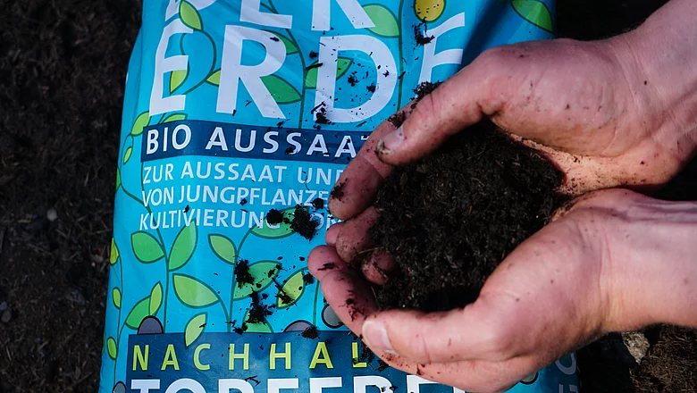 Nahaufnahme eines Paares von Händen, die Erde halten, im Hintergrund ist ein Beutel mit Dünger zu sehen